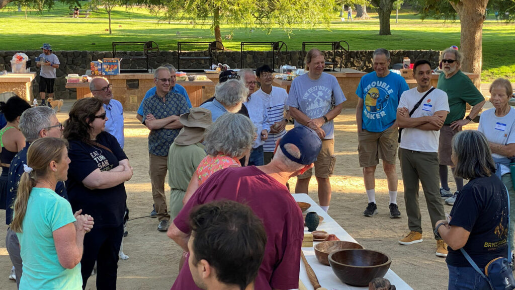 Members of woodturning clubs gather around a table displaying wooden bowls and projects during a joint picnic in Cuesta Park.