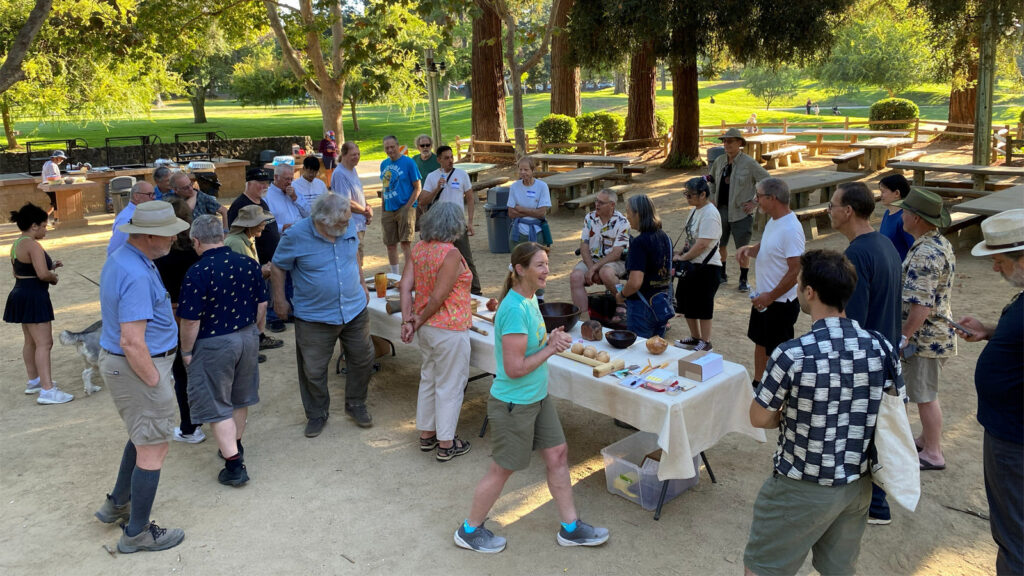 Members of the Silicon Valley and West Bay Woodturners gather around a table displaying turned wooden bowls and projects at an outdoor picnic in Cuesta Park, while a woman in a teal shirt speaks to the group.