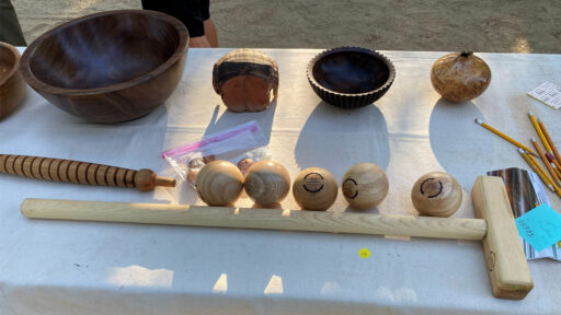 A table displays various woodturning projects, including a large dark bowl, a ribbed bowl, a burl wood vessel, a spiral-turned rod, and a wooden mallet, set out at the Silicon Valley and West Bay Woodturners joint picnic.