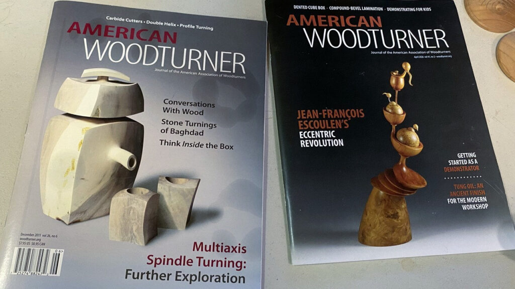Two issues of American Woodturner magazine featuring multi-axis turning lie on a table at a West Bay Woodturners meeting, with a blurred wooden project in the background.