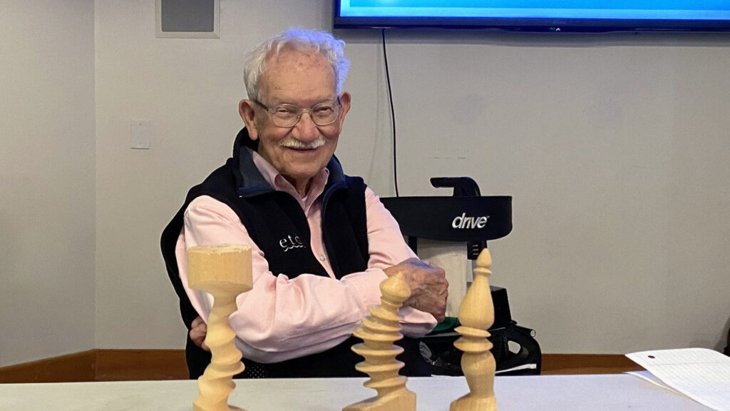 Bud Trapp poses with his multi-axis woodturning projects at a West Bay Woodturners meeting. Three light-colored wooden pieces with spiral designs sit on the table in front of him.