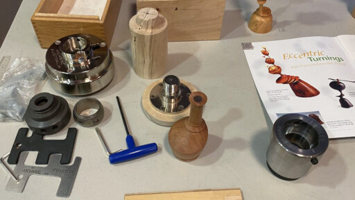 Specialized multi-axis chucks, a turned wooden vessel, and an instructional booklet on a display table demonstrating offset woodturning techniques at a West Bay Woodturners meeting.