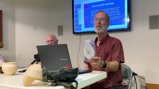 Dean Caudle demonstrates respiratory protection gear during a safety presentation at a West Bay Woodturners meeting, holding a mask with a health warnings slide displayed on a screen behind him.