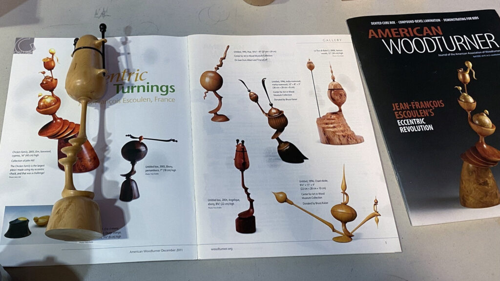 A display of Jean-Francois Escoulen's wooden sculptures and an open American Woodturner magazine at a West Bay Woodturners meeting, illustrating multi-axis turning techniques.