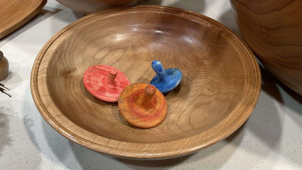 A shallow maple bowl crafted by Kelly Smith holds three colorful wooden spinning tops on a display table at a West Bay Woodturning meeting.