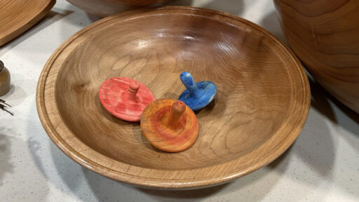 A shallow maple bowl crafted by Kelly Smith holds three colorful wooden spinning tops on a display table at a West Bay Woodturning meeting.