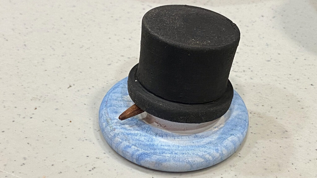 Melting Snowman woodturning sculpture by Kelly Smith, featuring a black top hat, small brown nose, and blue swirled base on a speckled white table.