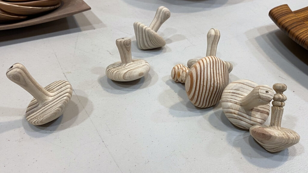Hand-turned wooden spinning tops by Kelly Smith, featuring various carved designs, displayed on a table at a West Bay Woodturners meeting in Los Altos, California.