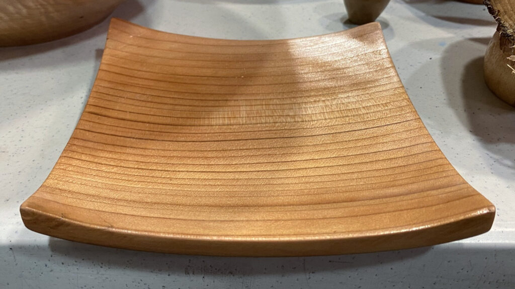 Golden-brown square woodturning plate with a grain texture by Kelly Smith. It sits on a white, speckled table at a West Bay Woodturners meeting.