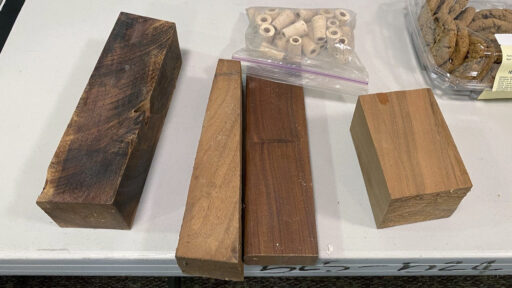 Participation prizes at a West Bay Woodturners meeting featuring raw wood blanks of various species, cork bottle stoppers, and a tray of cookies.