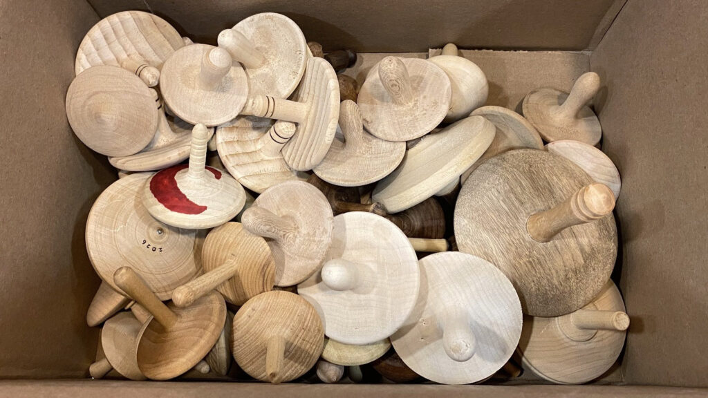 Assorted hand-turned wooden spinning tops in a cardboard box, donated by West Bay Woodturners for pediatric sickle cell anemia patients to demonstrate blood cell recovery.