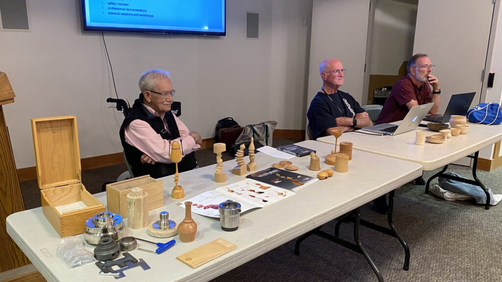 Bud Trapp, Edgar Whipple, and Dean Caudle sit behind a table laden with woodturning projects and tools at a West Bay Woodturners meeting in Los Altos, California.
